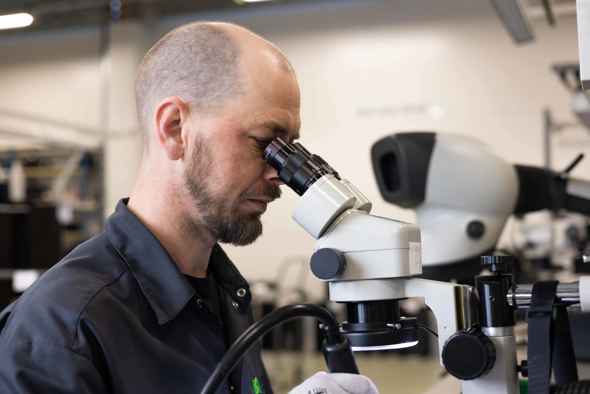 Sirico Electronicsin asiantuntija tarkastelee elektroniikkaa mikroskoopilla kuvastaen yrityksen tarkkuutta ja vastuullisuutta.